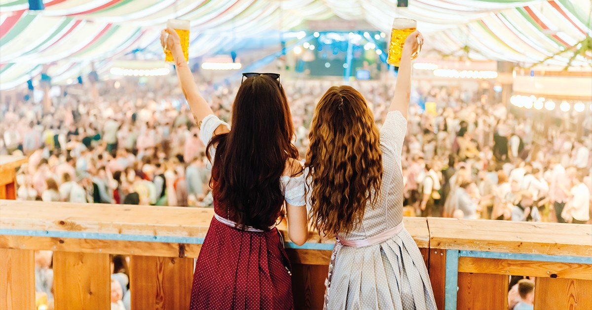 Duas visitantes em dirndl brindam sobre a plateia de uma tenda